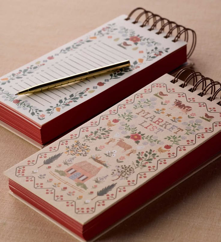 Two spiral-bound notepads with red edges and floral illustrations, one open with lined pages and a gold pen resting on top, and one closed showing a market list cover design, on a beige textured background.