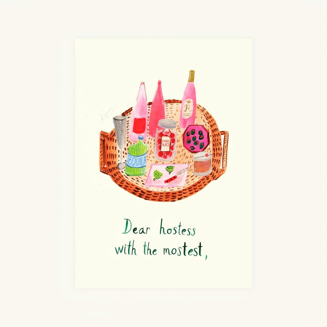 A square art print featuring a whimsical watercolor illustration of a wicker tray filled with pink bottles, a green hat, and a purple patterned cupcake, with the text 'Dear hostess with the most,!' below on a cream background.