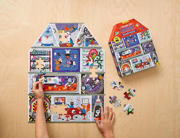 A children's jigsaw puzzle set displayed on a light wood surface, with a large assembled house-shaped puzzle, the retail box, and several loose pieces. A child's hands are visible interacting with the puzzle.