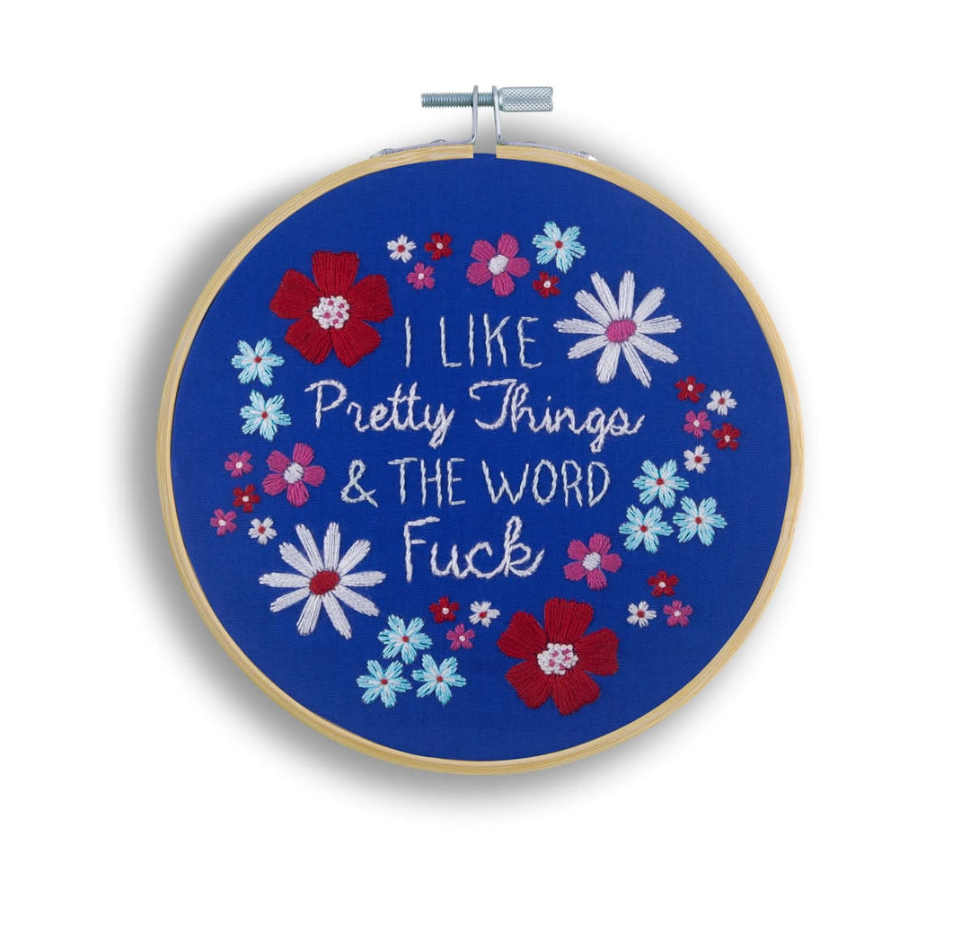 A circular embroidery hoop with a wooden frame and metal tension clamp, featuring a blue fabric design with embroidered white script text reading 'I LIKE Pretty Things & THE WORD Fuck' surrounded by red, pink, light blue, and white flowers.