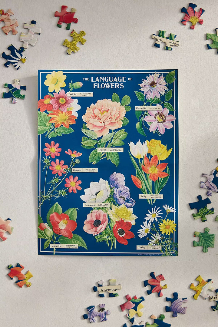 A vintage-style illustrated poster titled 'The Language of Flowers' laid flat on a light surface, surrounded by scattered jigsaw puzzle pieces. The poster features a deep blue background with colorful floral drawings and small text labels.