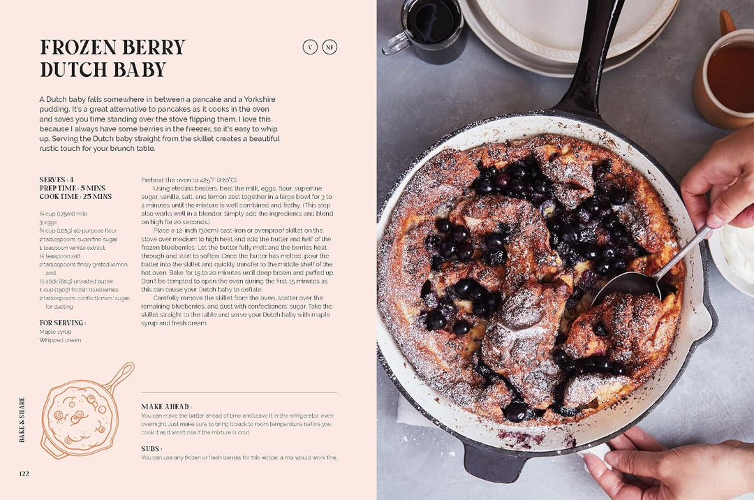 An open cookbook spread showing the recipe for Frozen Berry Dutch Baby on the left page and a photo of the prepared pancake in a cast-iron skillet on the right page, with hands holding the skillet.
