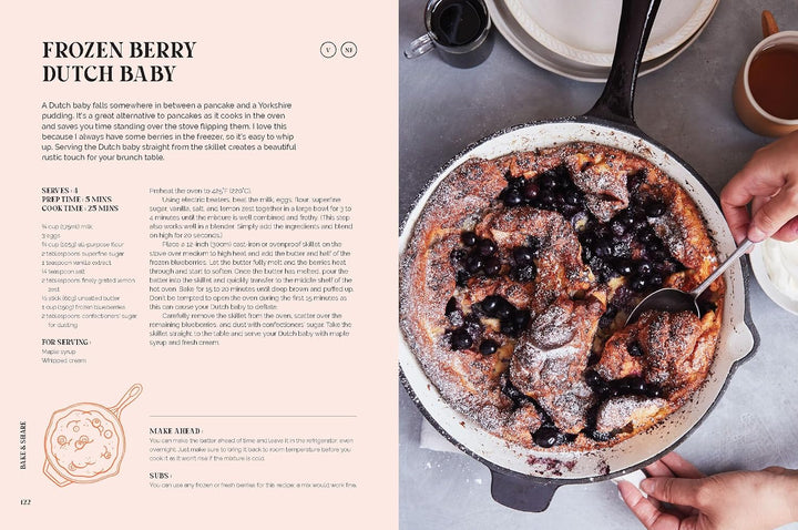 An open cookbook spread showing the recipe for Frozen Berry Dutch Baby on the left page and a photo of the prepared pancake in a cast-iron skillet on the right page, with hands holding the skillet.