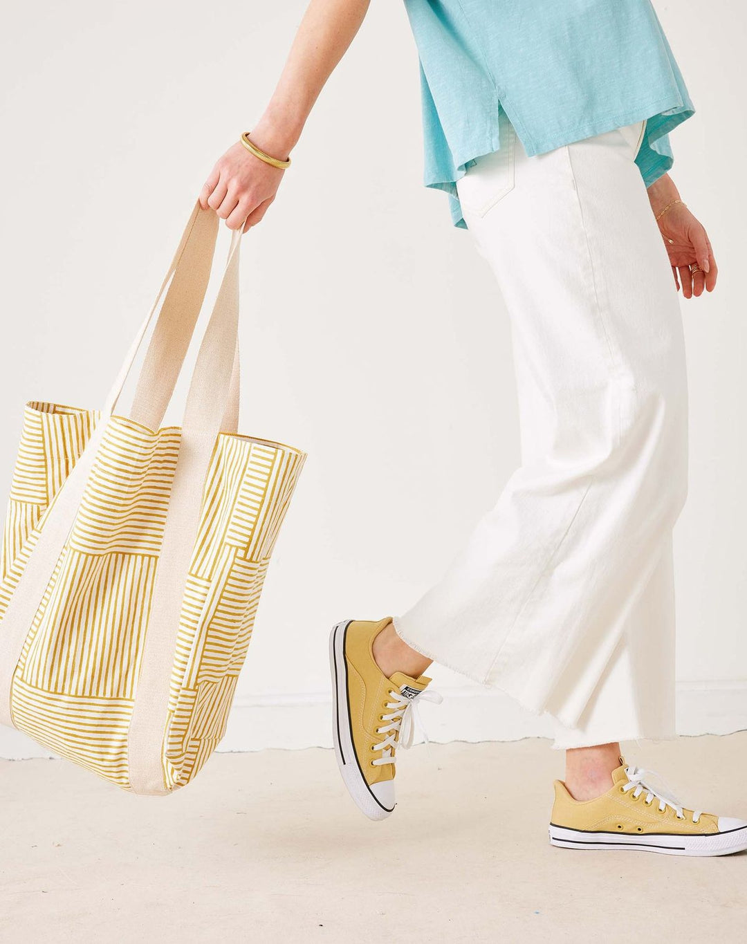 Person holding a striped tote bag wearing white pants and yellow sneakers on a plain background