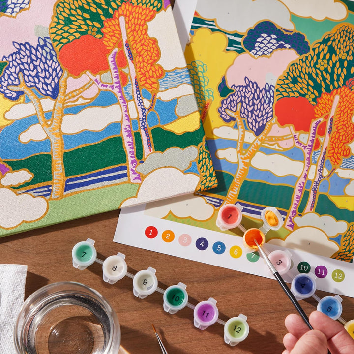 A hand is painting on a canvas using a paint-by-numbers guide and small paint pots on a wooden table. The partially painted canvas and guide show a colorful landscape with stylized trees, blue sky, green hills, and white clouds.
