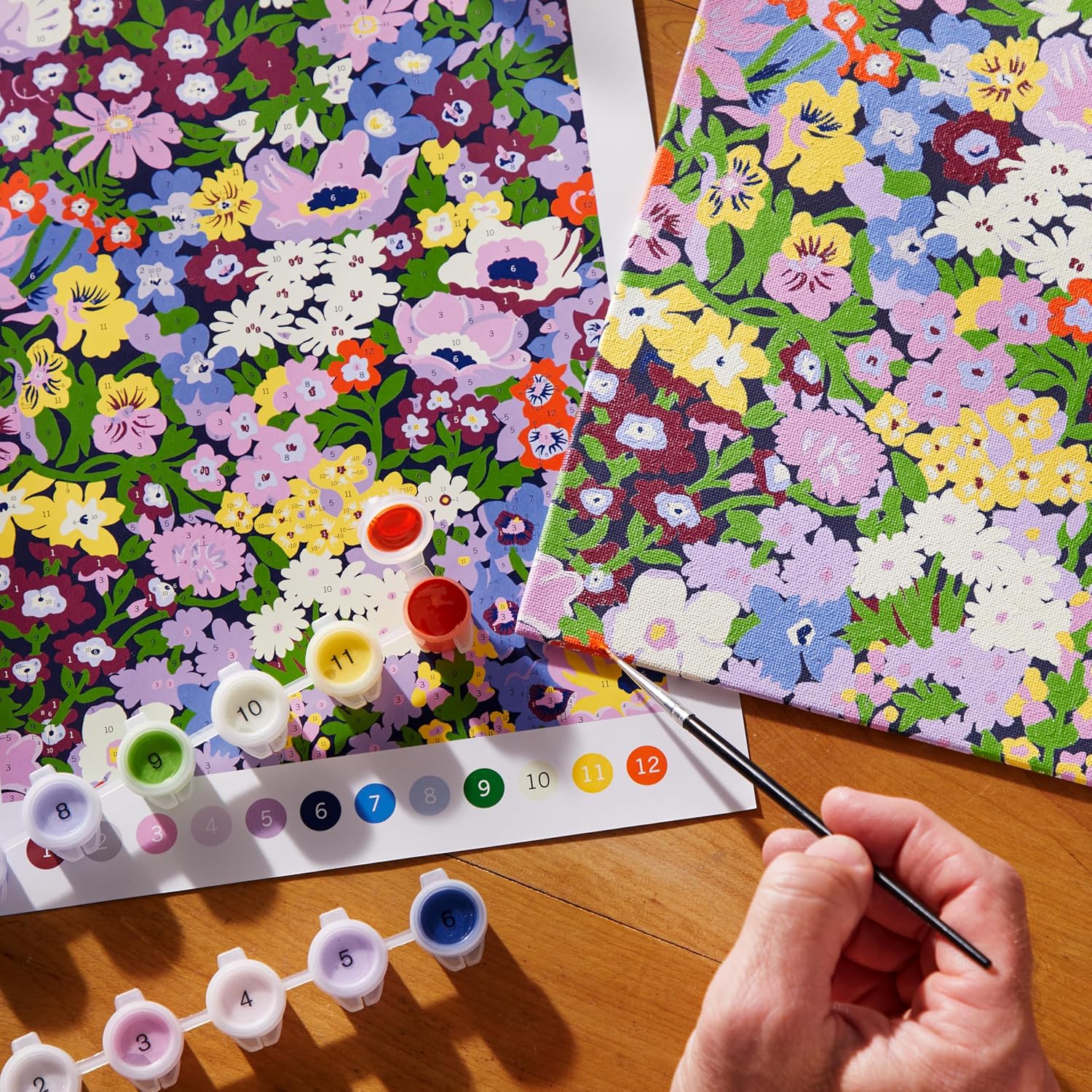 Floral patterned paper with paint swatches and a hand holding a paintbrush on a wooden surface.