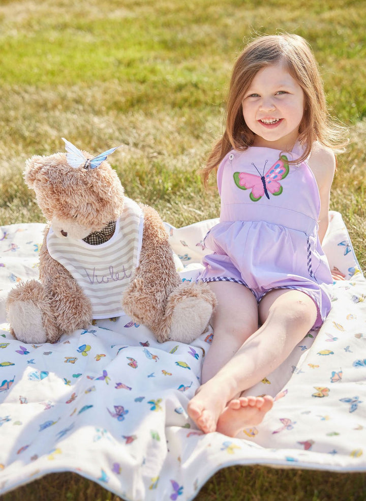 Young girl sitting on a blanket with a teddy bear, both on a grassy field