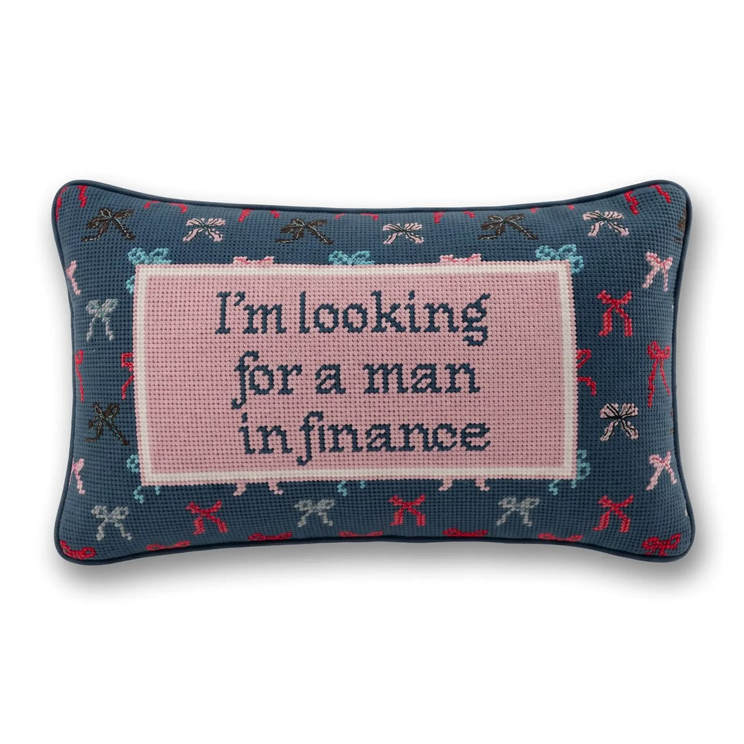 A rectangular decorative pillow with a navy blue base featuring a pattern of small bows in red, pink, and light blue. The center has a large light pink square with the text 'I'm looking for a man in finance' in dark blue lettering.