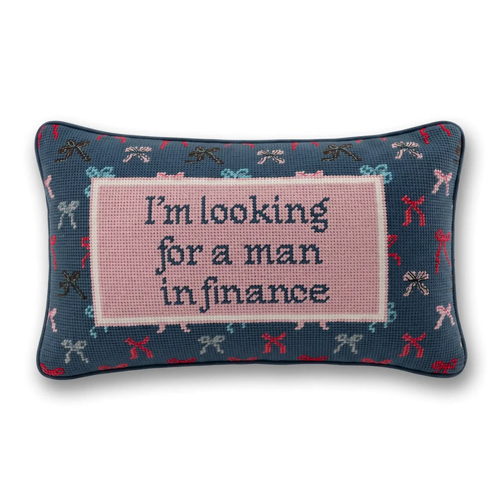 A rectangular decorative pillow with a navy blue base featuring a pattern of small bows in red, pink, and light blue. The center has a large light pink square with the text 'I'm looking for a man in finance' in dark blue lettering.