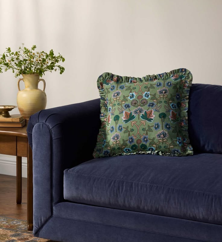 A decorative throw pillow with a green bohemian-style floral and bird pattern sits on a dark navy blue velvet sofa in a living room setting.