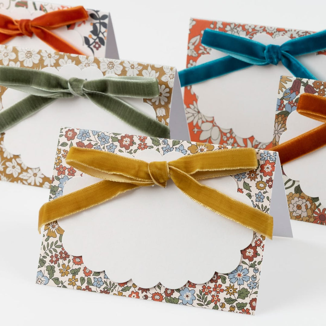 A set of floral patterned place cards with scalloped edges, each tied with a velvet ribbon bow in a different color including mustard yellow, teal, sage green, and rust orange, arranged on a white background.