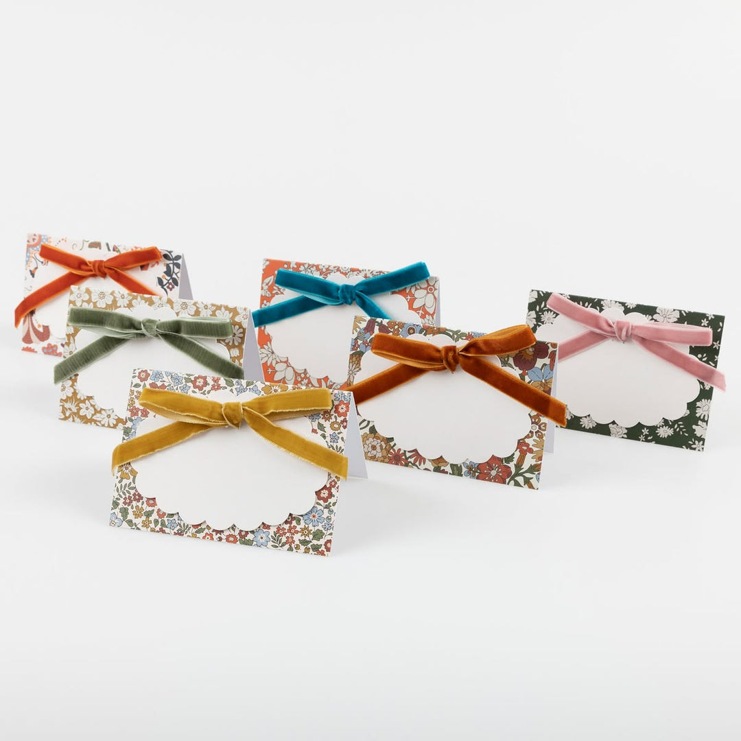 Five small floral place cards with white scalloped centers and tied velvet ribbons in orange, green, teal, mustard, and pink, arranged on a light gray background.