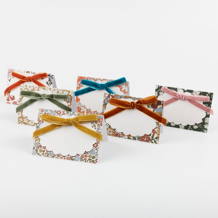 Five small floral place cards with white scalloped centers and tied velvet ribbons in orange, green, teal, mustard, and pink, arranged on a light gray background.
