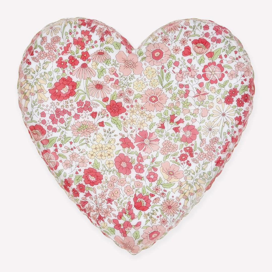 A heart-shaped decorative pillow featuring a dense, all-over floral print with pink, red, and yellow flowers on a white background, set against a solid light gray backdrop.