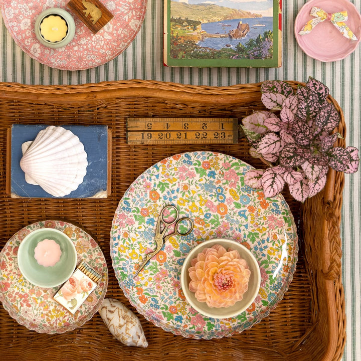 Decorative table setting with floral plates, a shell, and a book on a wicker tray.