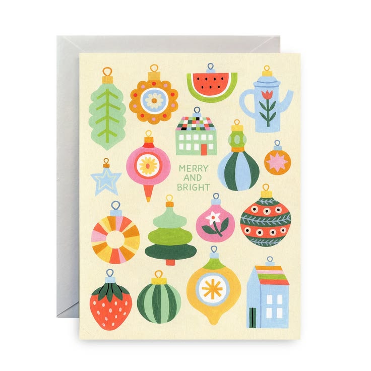 A Christmas greeting card with a light cream background, featuring colorful illustrated ornaments, a watermelon slice, a teapot, a strawberry, and the words 'MERRY AND BRIGHT' in the center. The card is shown standing upright with a gray envelope behind it on a white background.