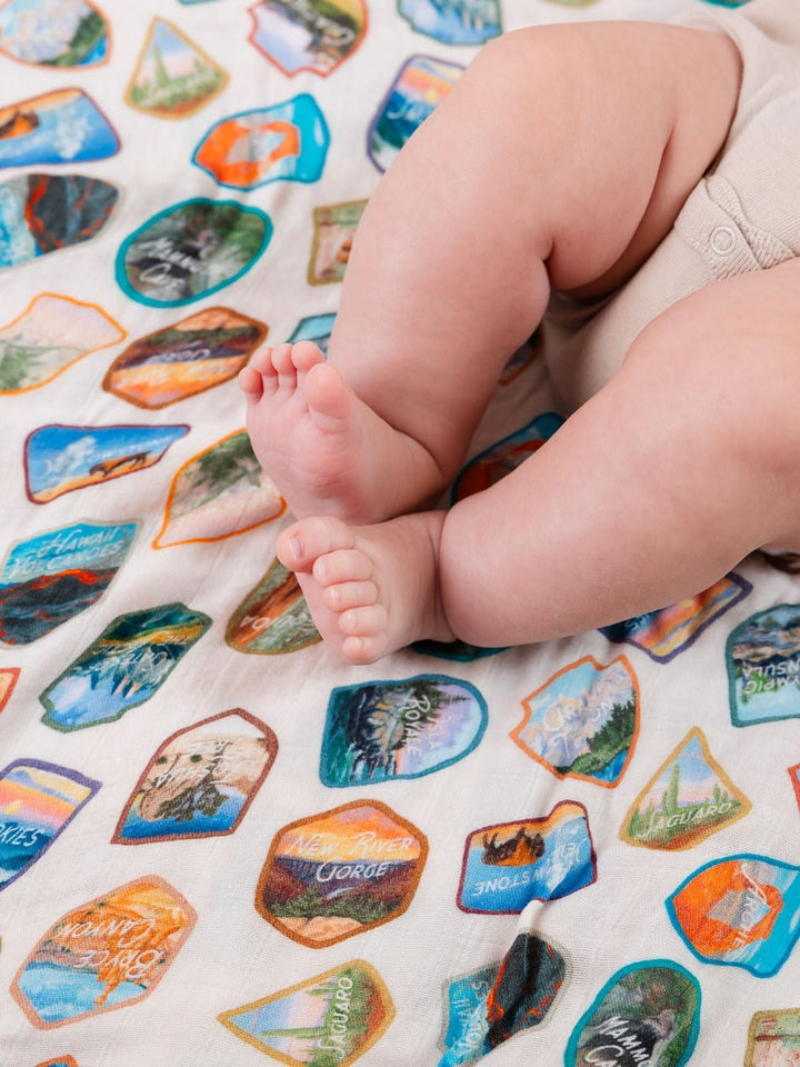 A close-up of a baby's legs and feet resting on a cream-colored blanket printed with colorful outdoor trail badge designs.