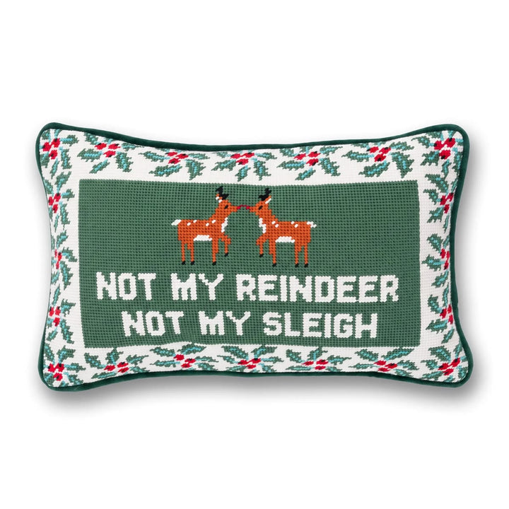 A rectangular Christmas needlepoint pillow with a dark green center featuring two orange reindeer and the text 'NOT MY REINDEER NOT MY SLEIGH' in white. The border is decorated with a white background, green holly leaves, and red berries, finished with a dark green stitched edge.