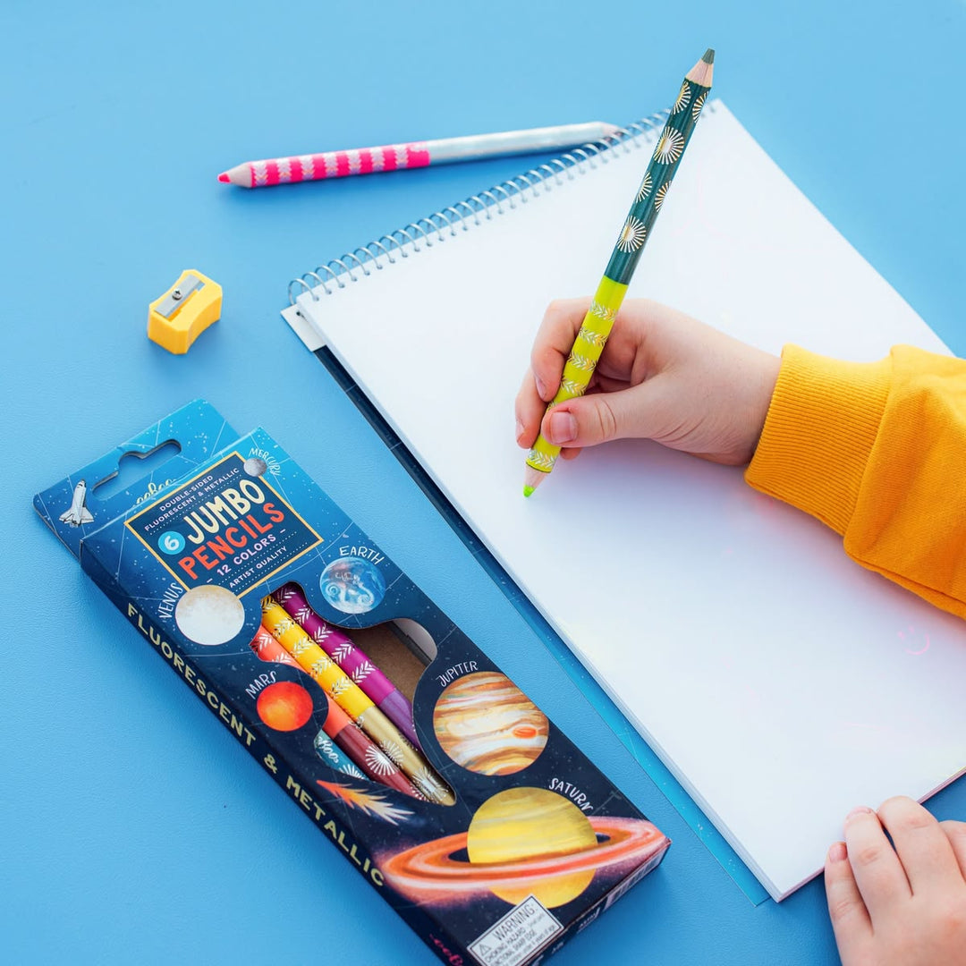 Hand holding a colorful pencil over a notebook with a package of Jumbo Pencils on a blue background