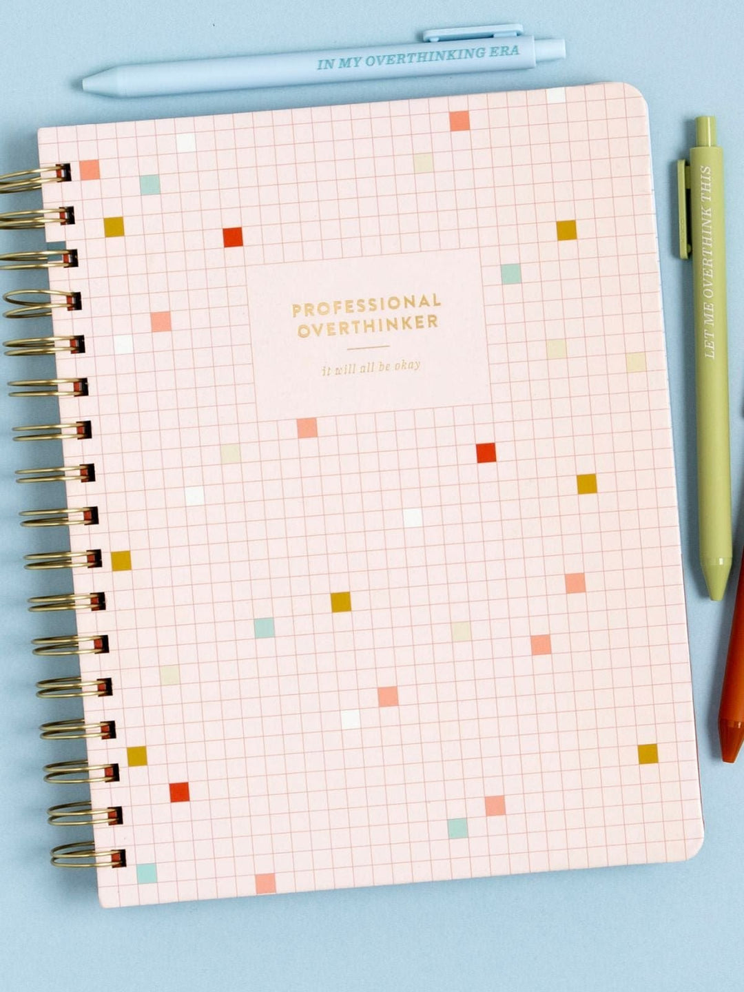 A spiral-bound planner or notebook with a pale pink grid cover, scattered small colored square markers, and the words 'PROFESSIONAL OVERTHINKER' on the front. A light blue pen at the top reads 'IN MY OVERTHINKING ERA,' and two green/orange pens sit to the right on a light blue background.
