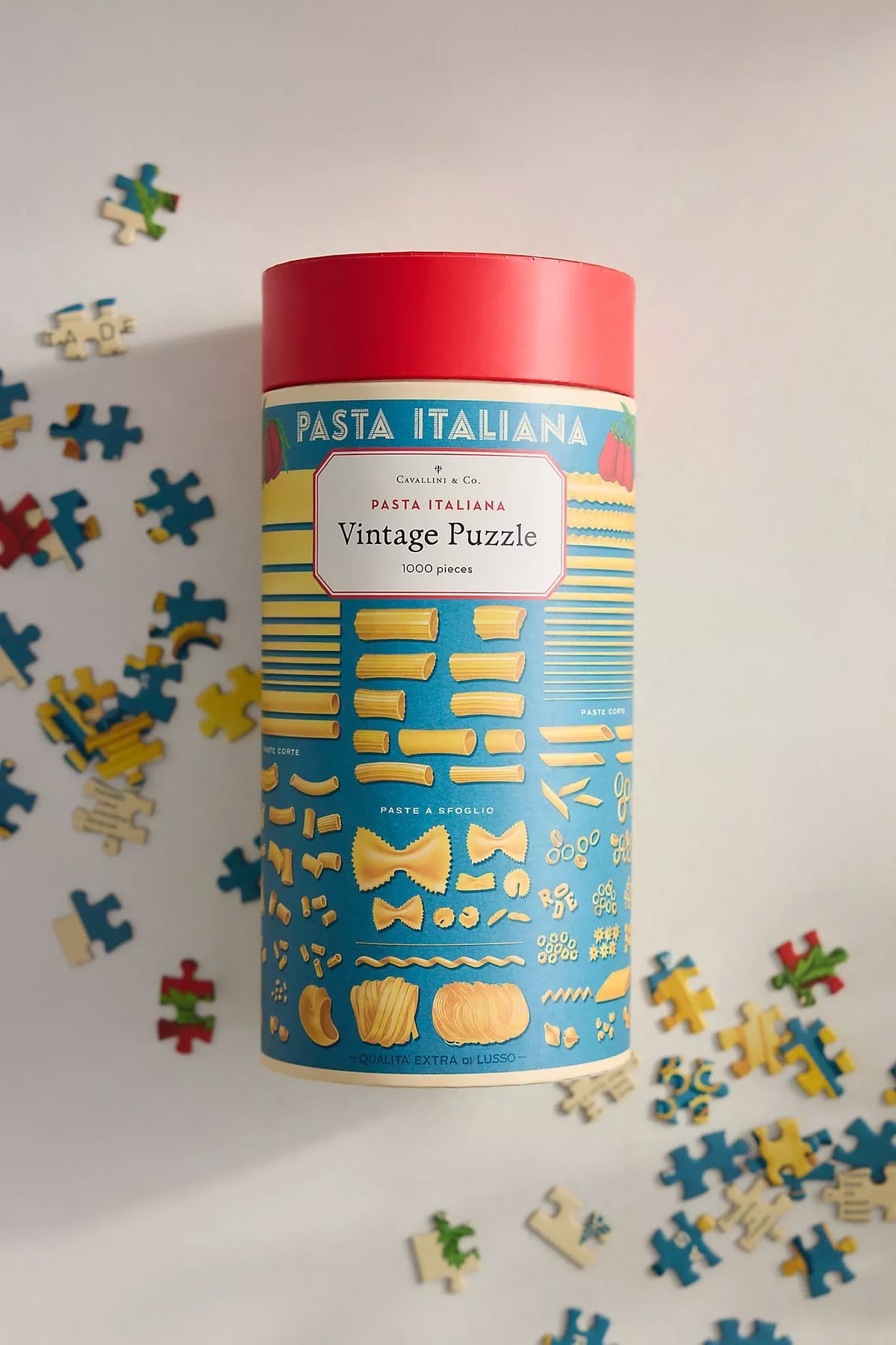 A cylindrical tin puzzle container labeled 'Pasta Italiana Vintage Puzzle' by Cucina & Co, with a red lid and a blue label printed with yellow pasta shapes, surrounded by scattered loose puzzle pieces on a white background.