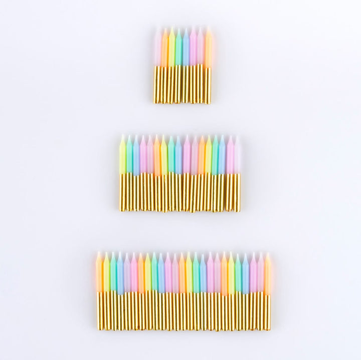 Three sets of colorful birthday candles arranged vertically on a plain white background. The candles have pastel pink, yellow, green, blue, and orange tips with shiny gold wax bases.