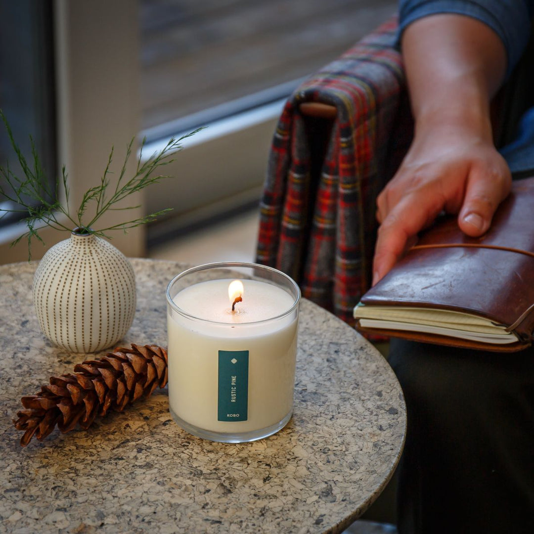 Candle with a label on a table next to a pine cone and small vase, with a person holding a book in the background.
