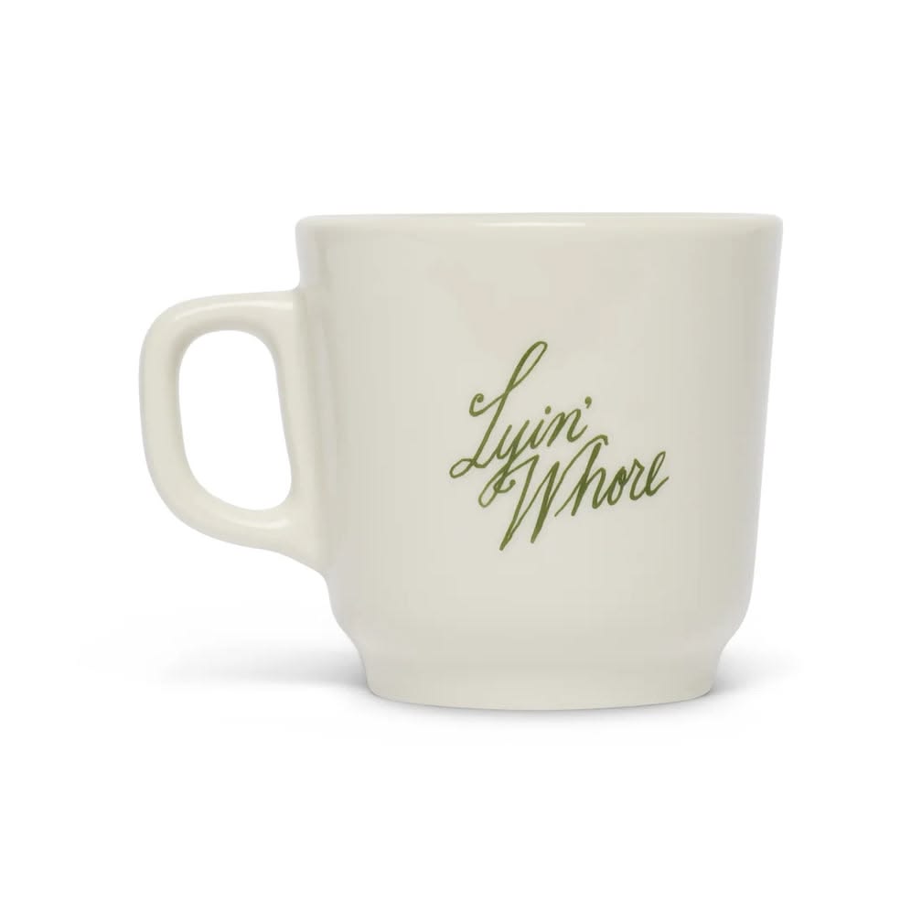 An off-white ceramic mug with a green cursive script reading 'Lyin' Whore' on the side, set against a plain white background.