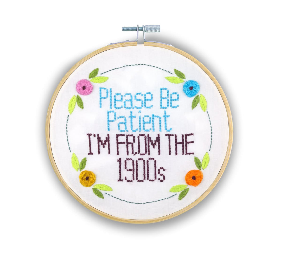 A round embroidery hoop containing a white fabric with colorful cross-stitch text that reads 'Please Be Patient I'M FROM THE 1900s' surrounded by a floral wreath design, shown on a white background.