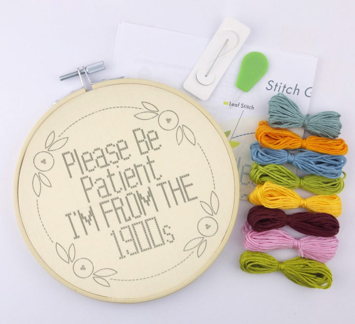Cross-stitch kit with a wooden hoop showing the finished design reading 'Please Be Patient I'M FROM THE 1900s', plus thread skeins in multiple colors, a green leaf-shaped tool, and a white clip on a white background.
