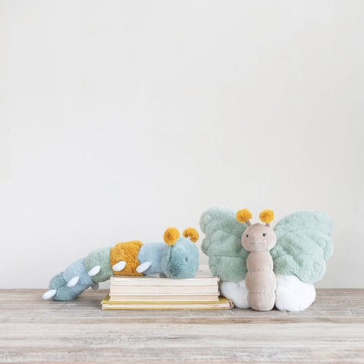 Two plush caterpillar toys arranged on a light wood surface against a plain off-white wall. One caterpillar lies on stacked books, and the other stands beside them.