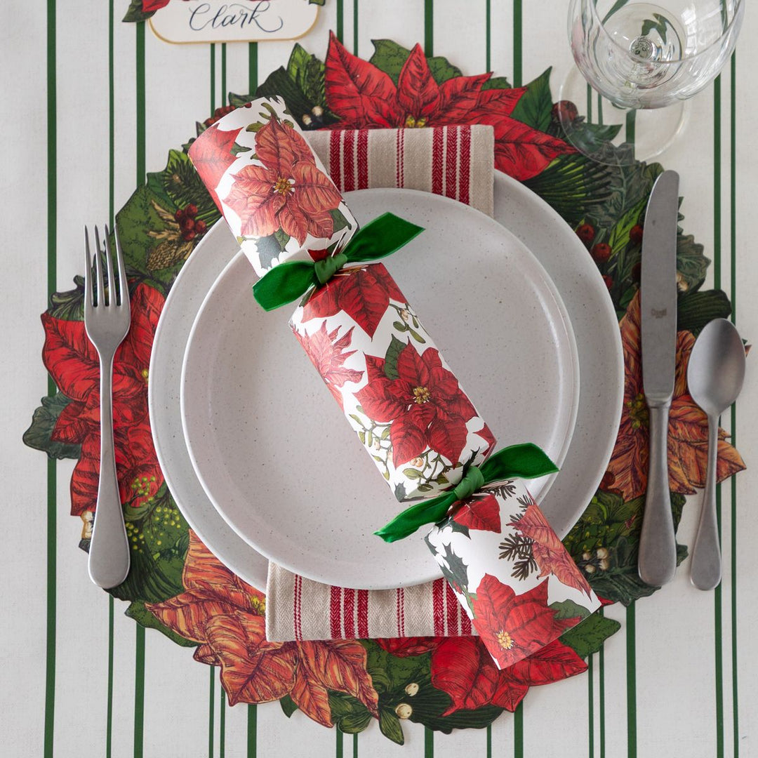 Christmas-themed table setting with poinsettia napkin, greenery placemat, and cutlery on a striped background.