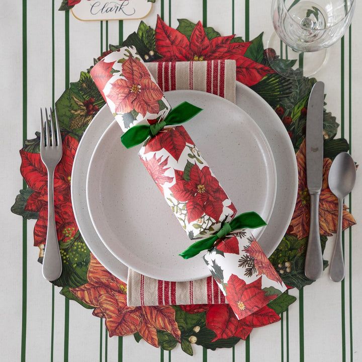 Christmas-themed table setting with poinsettia napkin, greenery placemat, and cutlery on a striped background.