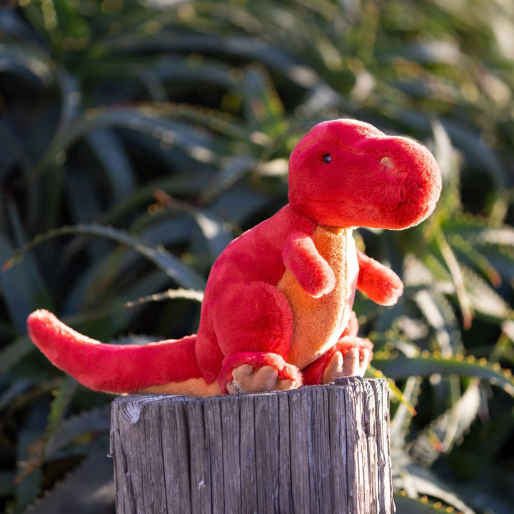 Red and orange plush dinosaur toy on a wooden post with green plants in the background