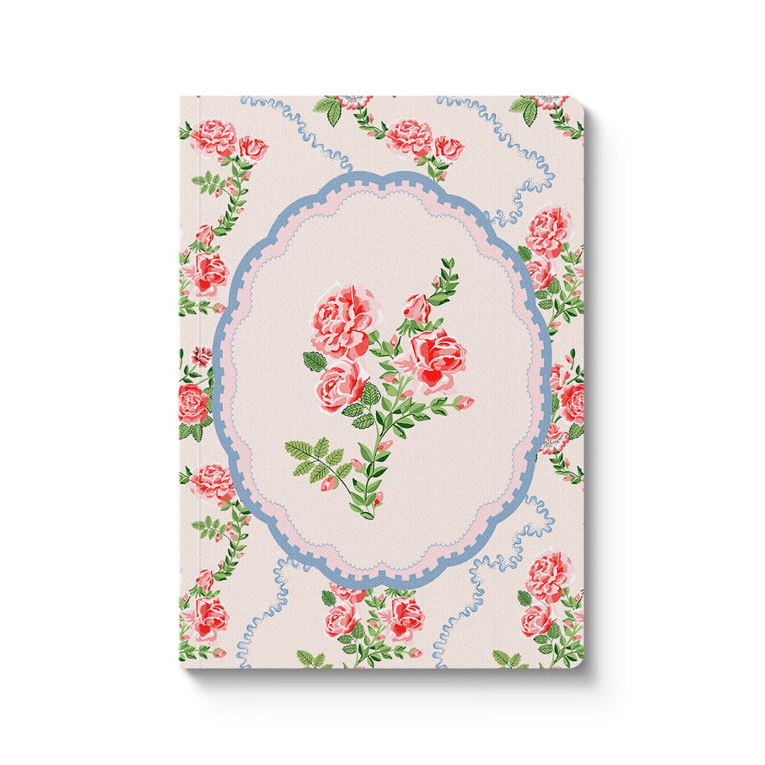 A notebook with a vintage floral cover design, featuring pink roses and green leaves inside a blue oval frame on a cream background, shown against a plain white background.