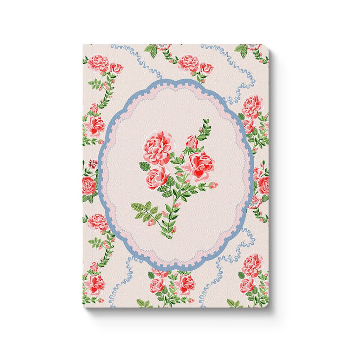 A notebook with a vintage floral cover design, featuring pink roses and green leaves inside a blue oval frame on a cream background, shown against a plain white background.