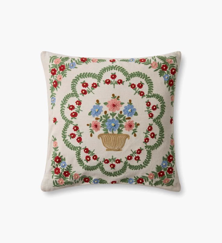 A square decorative throw pillow with a cream-colored linen background, featuring a central embroidered floral bouquet in a basket surrounded by a green leafy wreath and red, pink, and blue flowers.