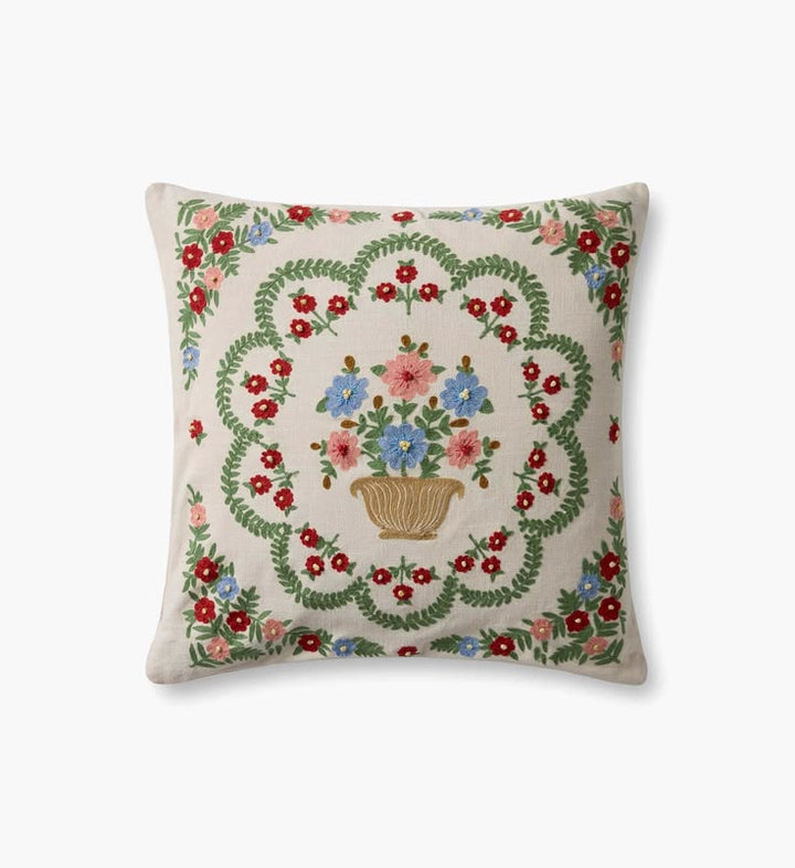 A square decorative throw pillow with a cream-colored linen background, featuring a central embroidered floral bouquet in a basket surrounded by a green leafy wreath and red, pink, and blue flowers.