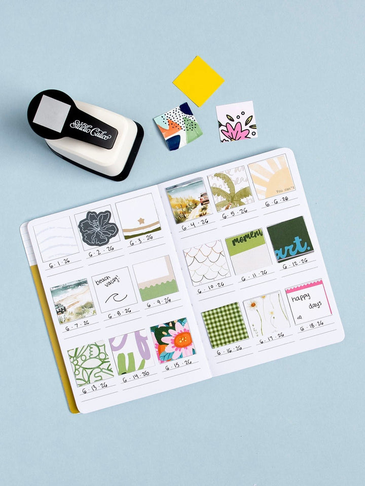 An open planner or scrapbook journal laid on a light blue background, filled with small decorative paper inserts and cards. A black-and-white die-cut hole punch sits at the upper left, with a few loose patterned paper pieces and a yellow sticky note above the journal.
