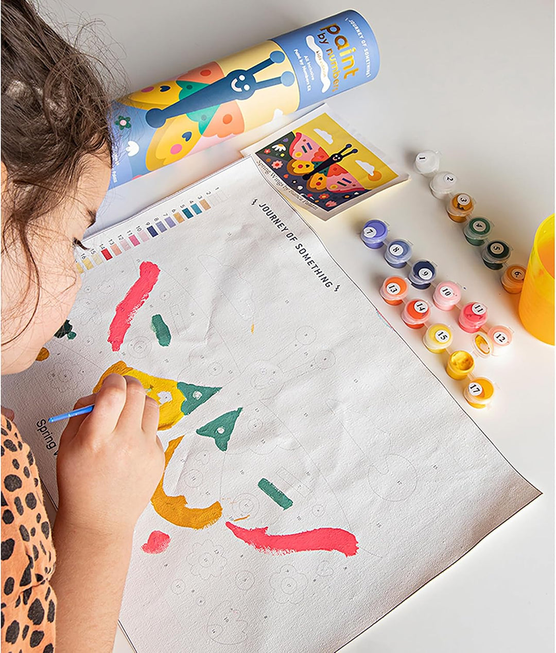 A child's hand and head are visible as they paint on a large sheet of paper. Surrounding the artwork are small colorful paint tubes, a paint palette, and a rolled tube of paint on a white tabletop.