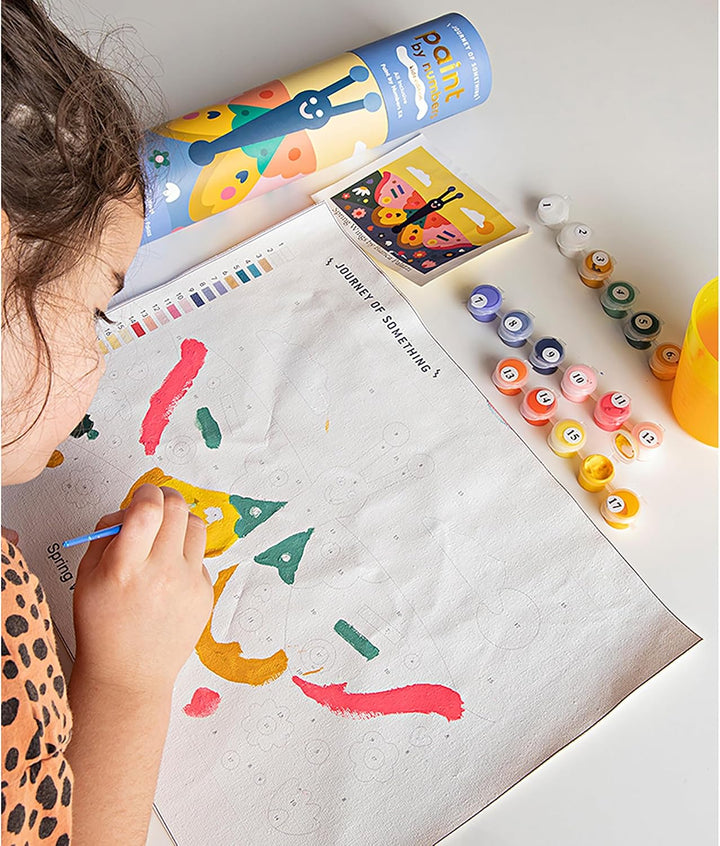 A child's hand and head are visible as they paint on a large sheet of paper. Surrounding the artwork are small colorful paint tubes, a paint palette, and a rolled tube of paint on a white tabletop.