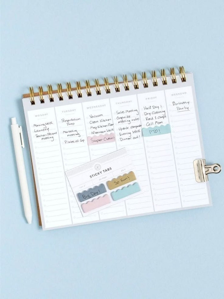 An open spiral-bound planner on a light blue background, showing weekly lined pages with handwritten notes and pastel sticky tabs. A white pen and a metal clip are placed beside it.