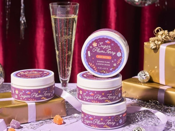 Jars of 'Sugar Plum Fairy' body scrub with a glass of champagne and gift boxes on a decorative surface.