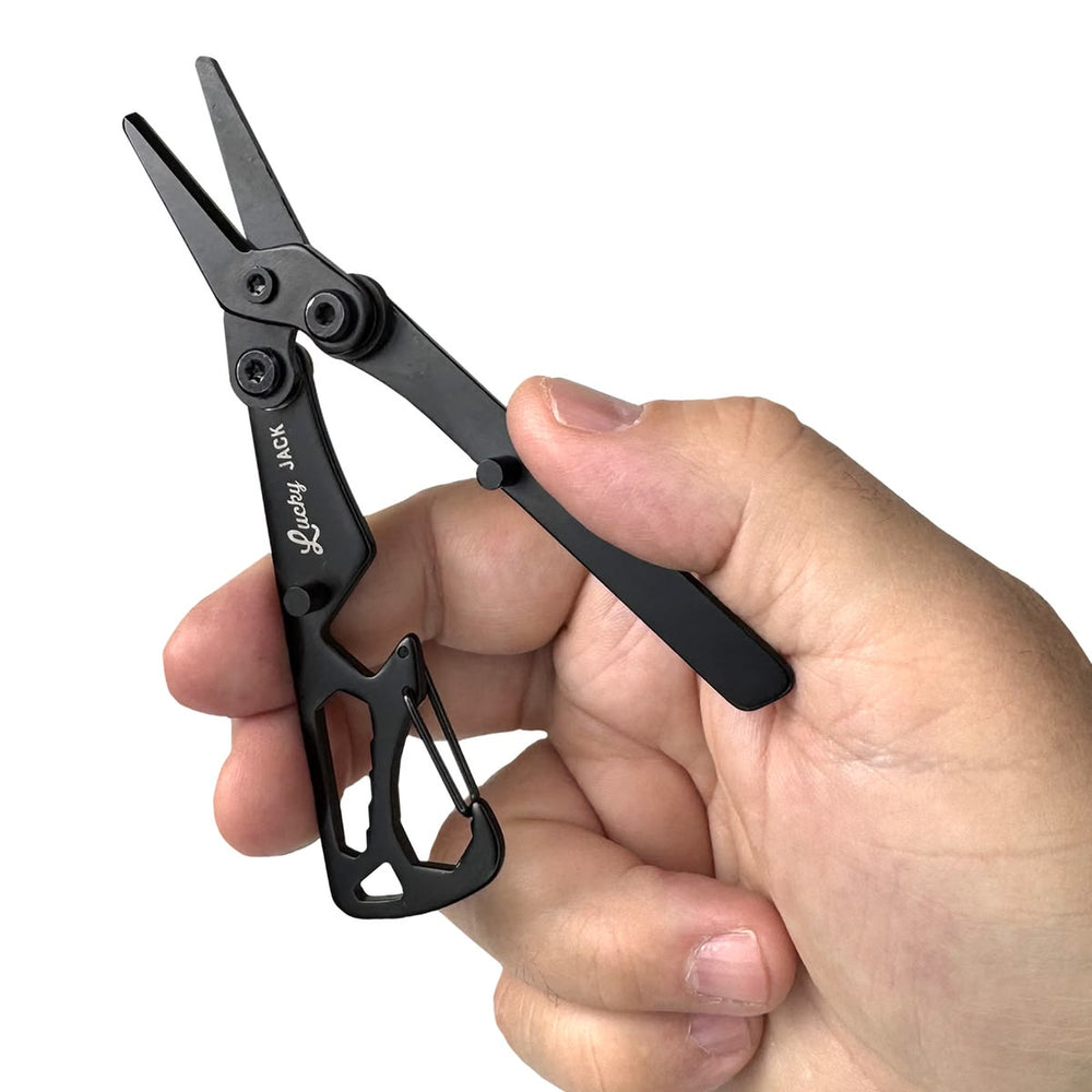 A hand holding a compact black metal multi-tool scissors with a carabiner clip, shown against a plain white background.