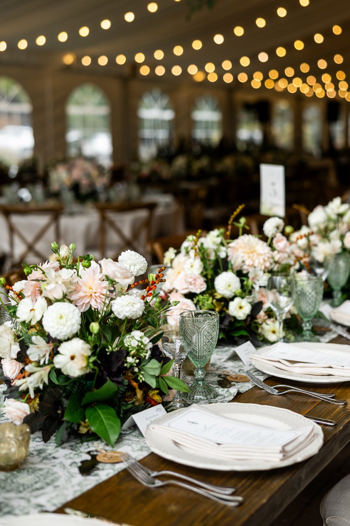 A styled wedding reception table setting with lush floral centerpieces in white, blush, and green, paired with white plates, folded napkins, and textured green glassware, under warm string lights in a large event hall.
