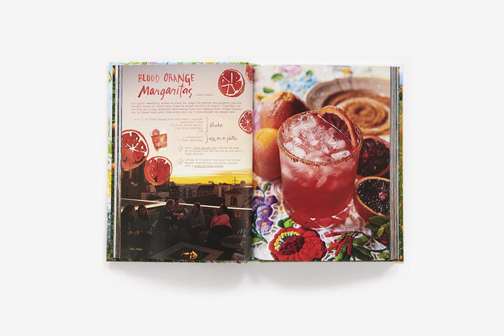 An open printed recipe book or magazine spread showing a cocktail recipe titled 'Blood Orange Margaritas,' with a photo of a pink drink on the right page and recipe text on the left, photographed on a light gray background.