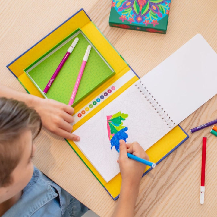 Child drawing with colored pencils on a notebook with a colorful box of pencils next to it.