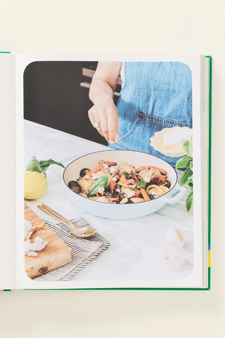 A hardcover cookbook or recipe book shown open on a light cream background. The cover features a large photograph of a person in a blue apron seasoning a bowl of pasta on a kitchen counter, with a lemon and wooden spoon nearby.