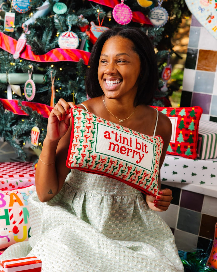 A woman smiling in a holiday-themed setting, holding a rectangular decorative pillow that reads 'a' tini bit merry' with a red and green Christmas pattern border. A decorated Christmas tree and wrapped gifts are visible in the background.