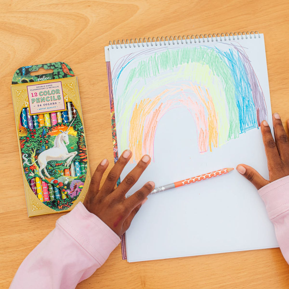 Child coloring a rainbow on a notebook with a box of color pencils on a wooden table